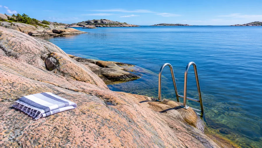 Slät rosa-grå granit som sluttar ner i klart blått hav, en metallbadstege fastsatt i berget
